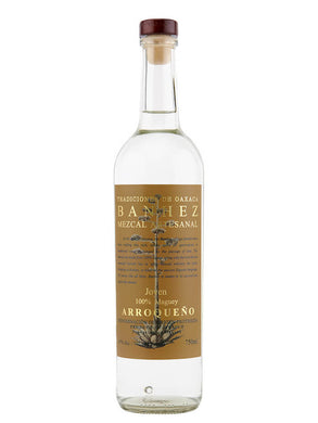 Banhez Mezcal Arroqueño bottle displayed on a rustic wooden table with a backdrop of agave plants under soft natural lighting, evoking a handcrafted, artisanal feel.